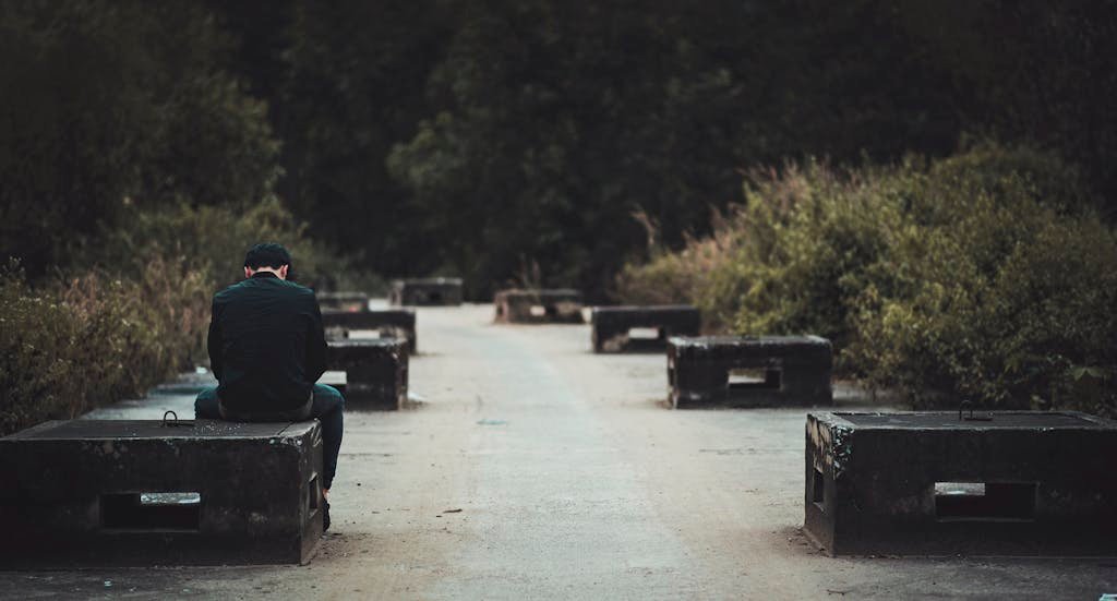 A man sitting alone on a bench along a concrete path in a serene outdoor setting.