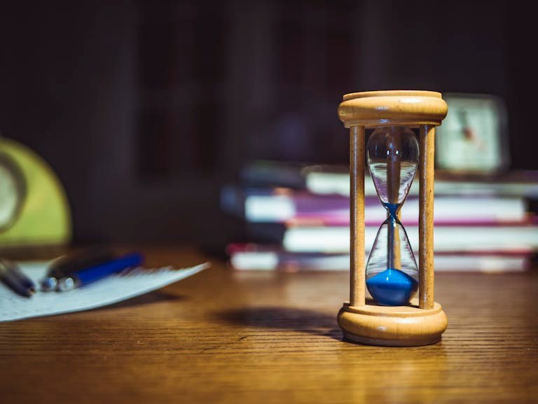 Close-up of a wooden hourglass on a desk, alongside books and pens, suggesting focused study or time management.
