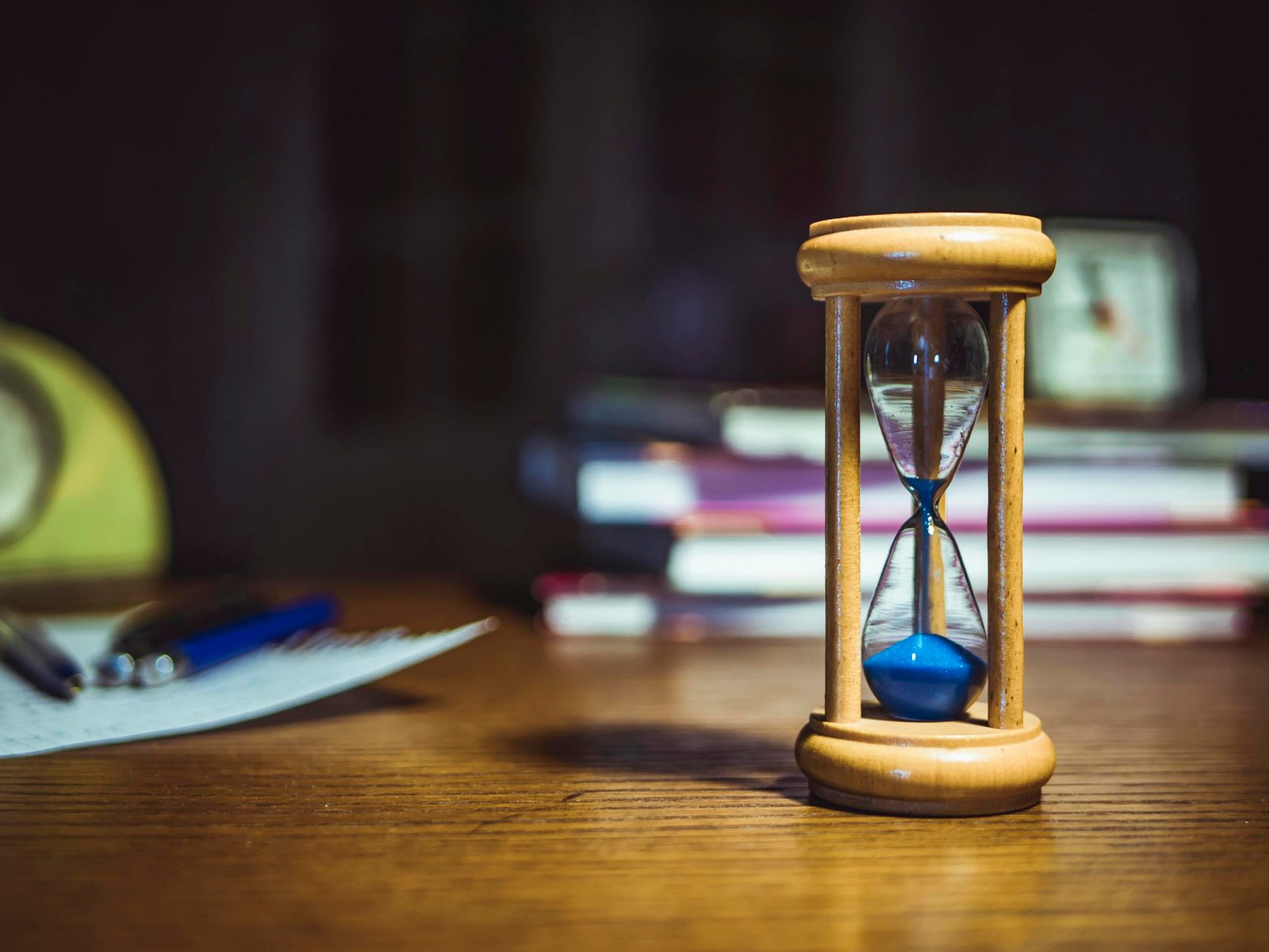 Close-up of a wooden hourglass on a desk, alongside books and pens, suggesting focused study or time management.
